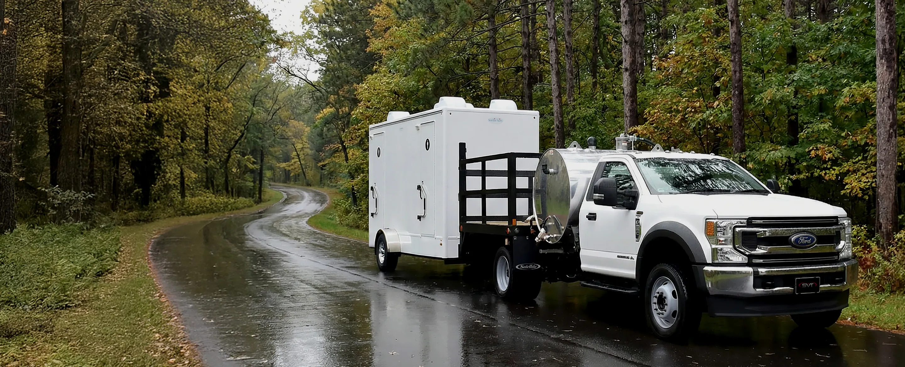 White Ford truck with a water tank and trailer parked on a wet road surrounded by trees.